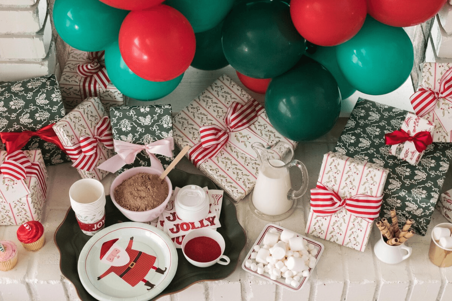 A crowded display of red and green wrapped Christmas presents and balloons with a tray of hot chocolate and marshmallows.