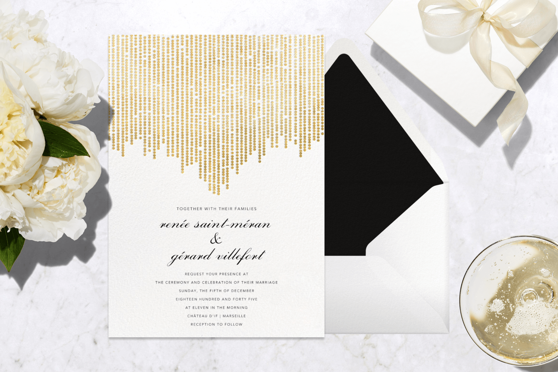 A white and gold wedding invitation surrounded by flowers, a present, and a glass of champagne.