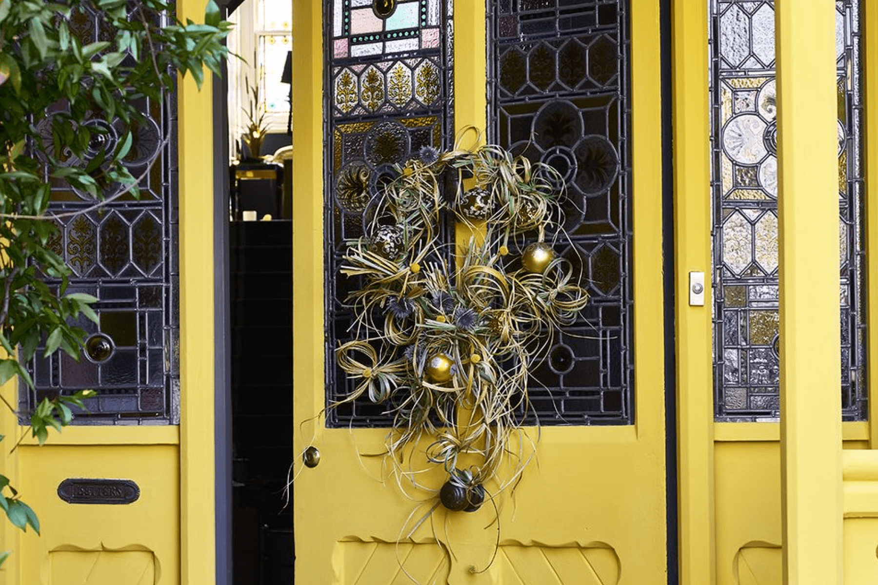 yellow door with stained glass windows and a modern wreath made of knotted fronds and gold Christmas tree ornaments is slightly ajar