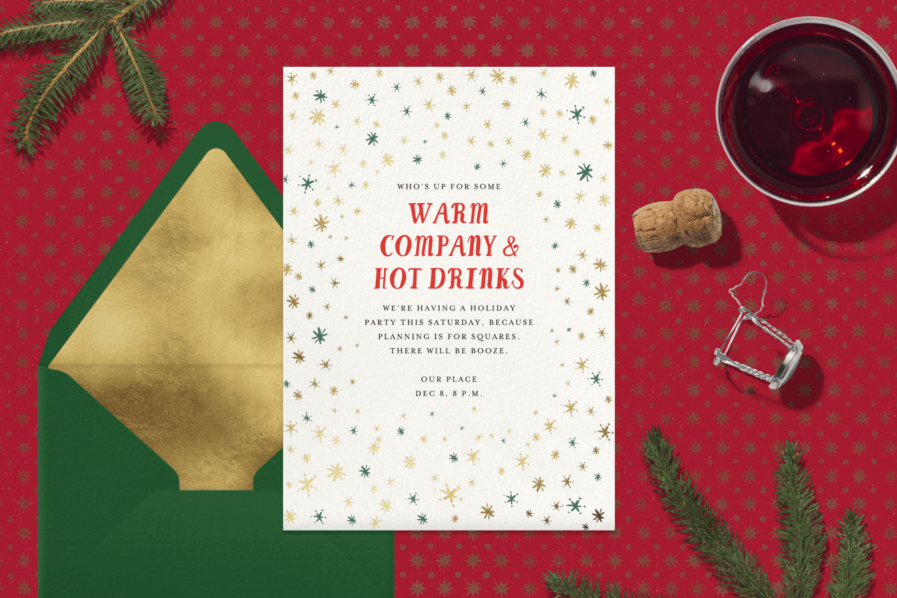 white invitation for “Warm company & hot drinks” with small gold and green asterisk stars lies on a red background beside a red drink, bottle cork, and green fir branches