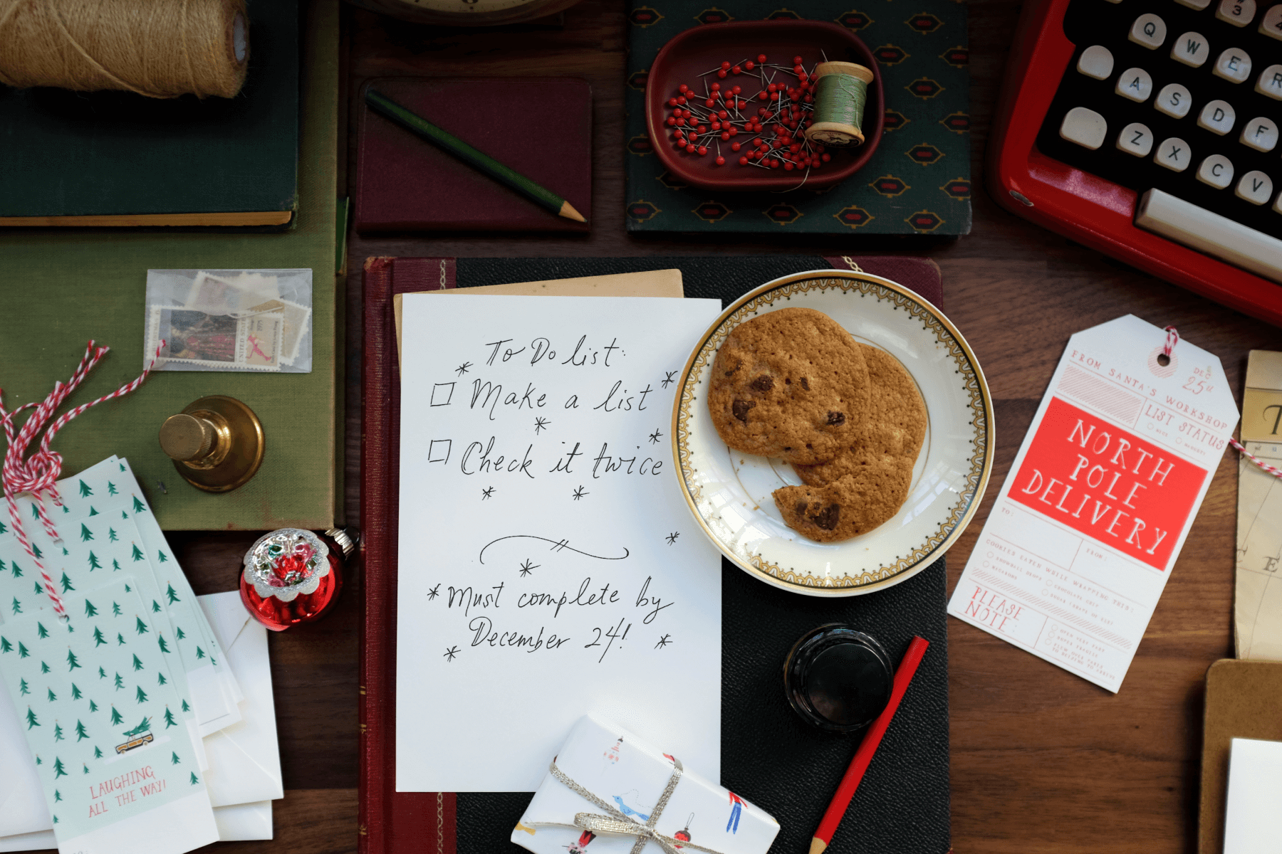 A to-do list reading “Make a list, check it twice” on what appears to be a desk belonging to Santa Claus, surrounded by cookies, a North Pole delivery tag, small wrapped gifts, and a red typewriter.