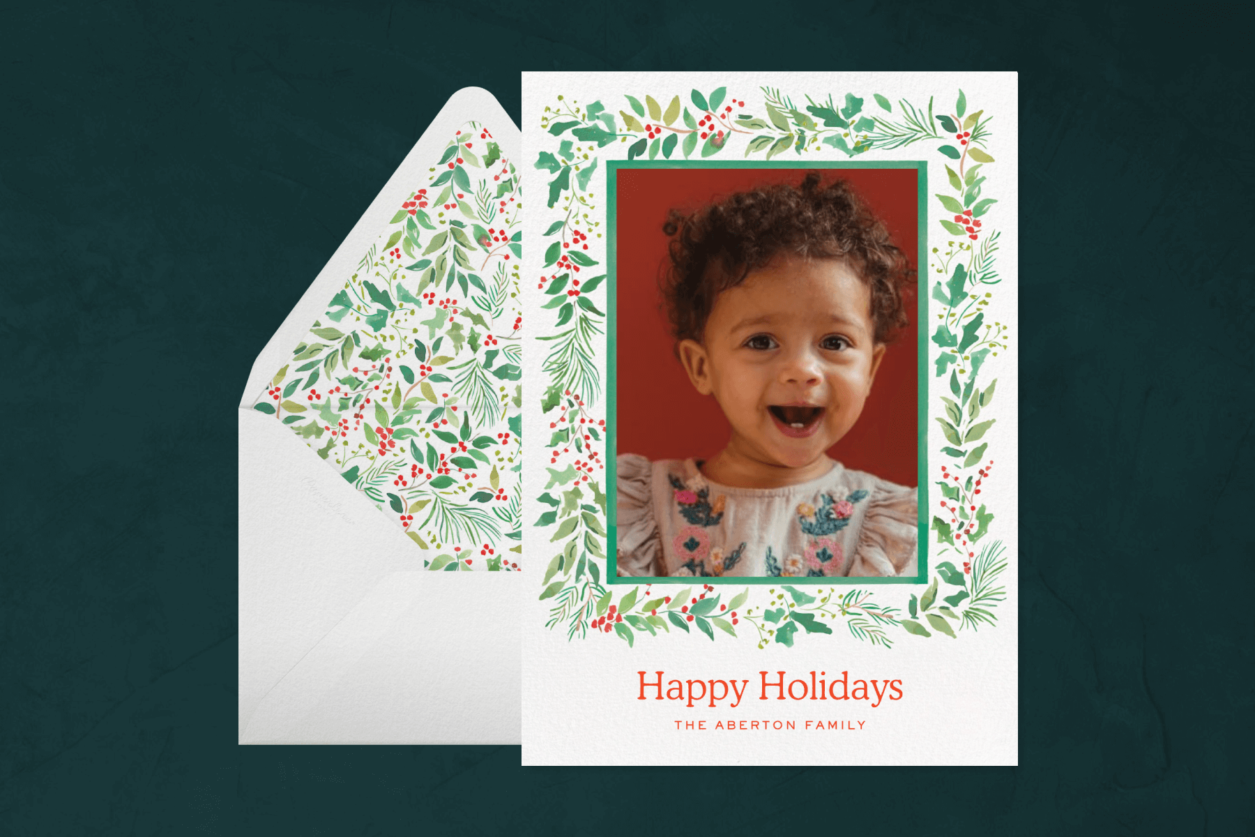 A holiday card featuring a baby photo in a garland frame, in front of an envelope with a matching garland liner