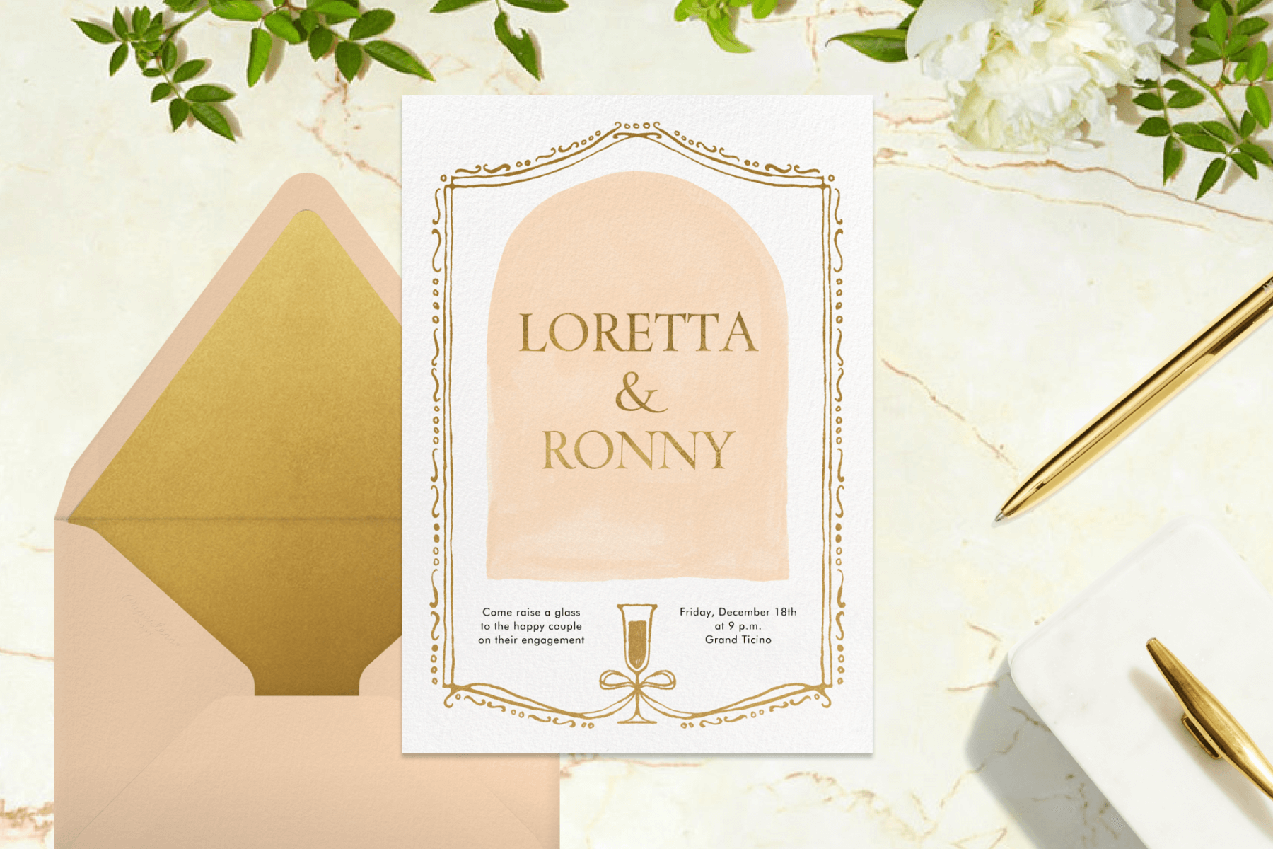An engagement party invitation with an arch-shaped text box and an ornate frame with a gold champagne glass. The invitation is paired with a pink and gold envelope and is surrounded by greenery, a gold pen, and a small box.