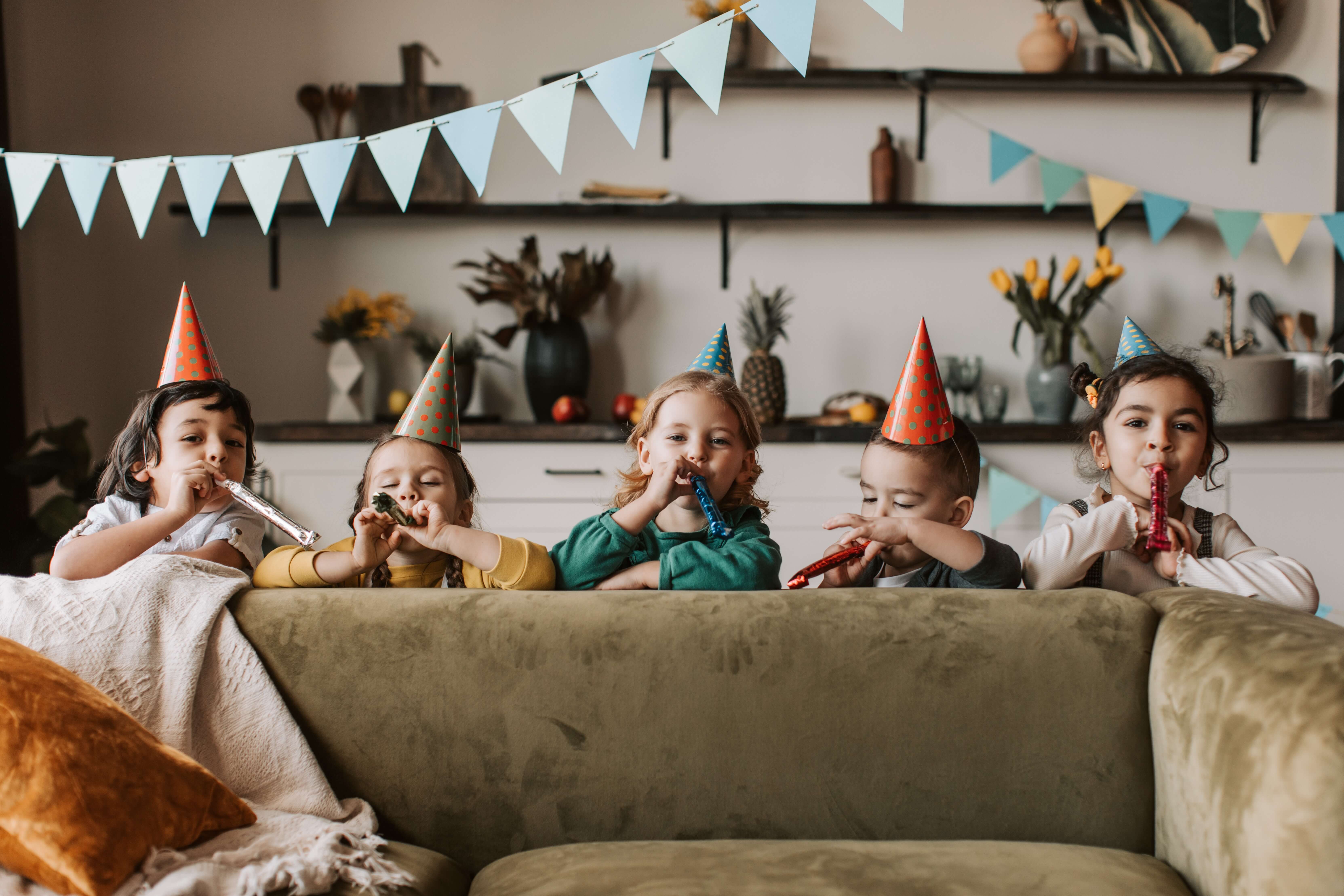Five children in party hats pop out from behind a couch and blow on noisemakers.