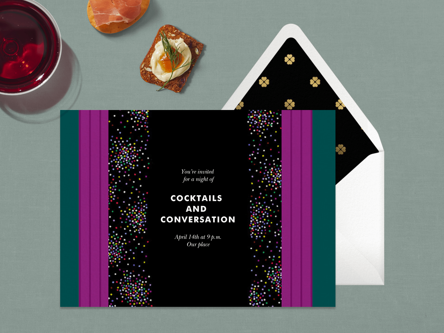 A cocktail party invitation with forest green, fuchsia, black, and rainbow confetti stripes next to a red drink and crackers.