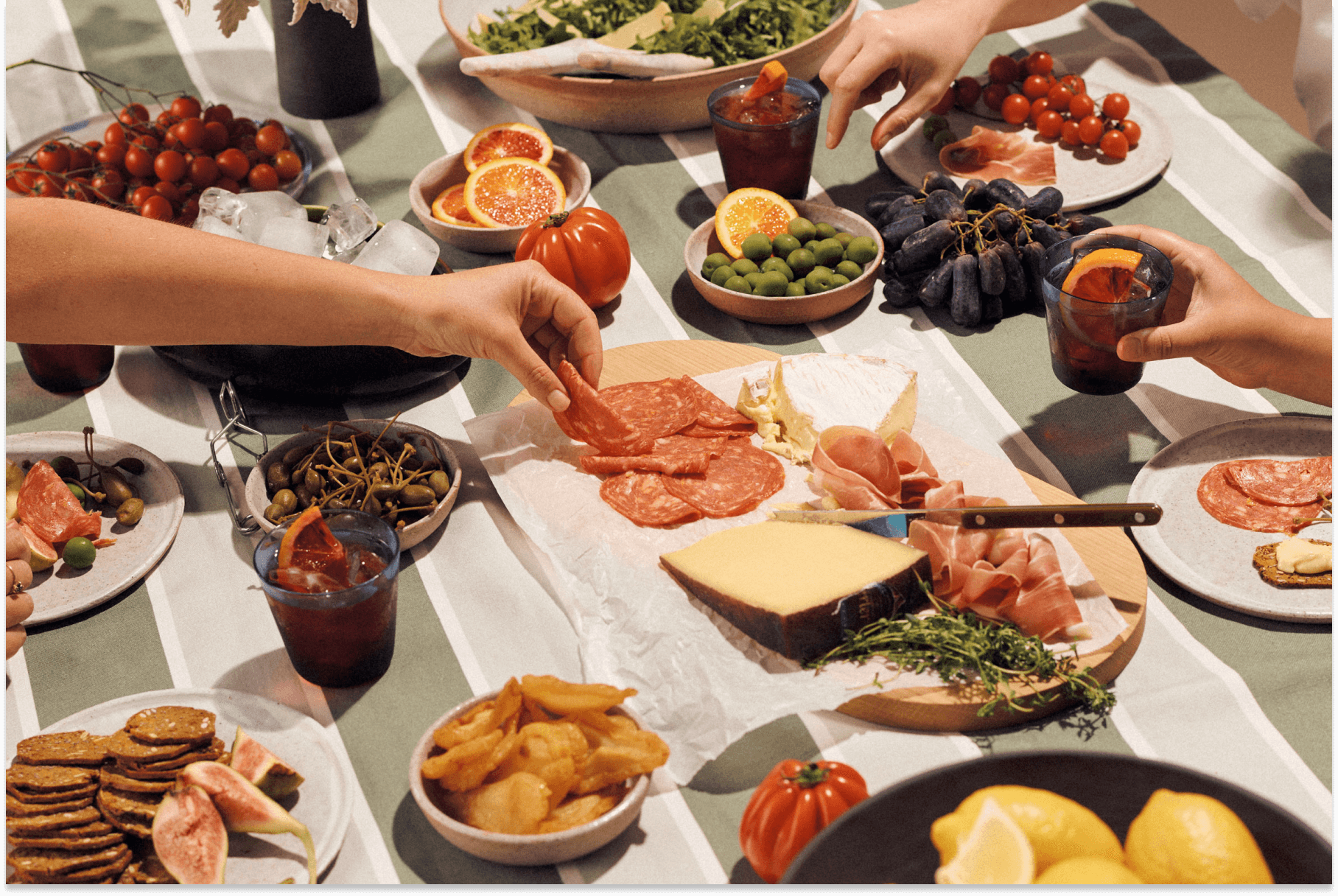 Hands reach over a green striped table top covered in finger food, like cheese and charcuterie, olives, grapes, cherry tomatoes, and more.