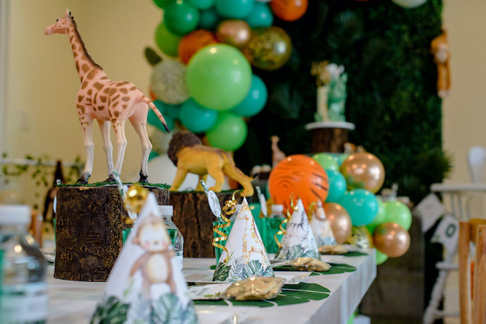 A table set for a children’s birthday party with giraffe and lion figurines as centerpieces, green and orange balloons, and party hats at each place setting.