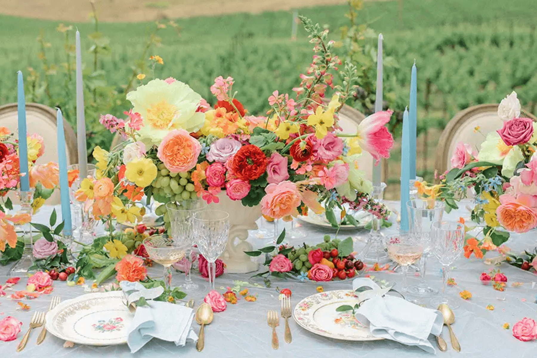 An outdoor table is set with colorful flowers spilling onto the place settings and light blue taper candles.