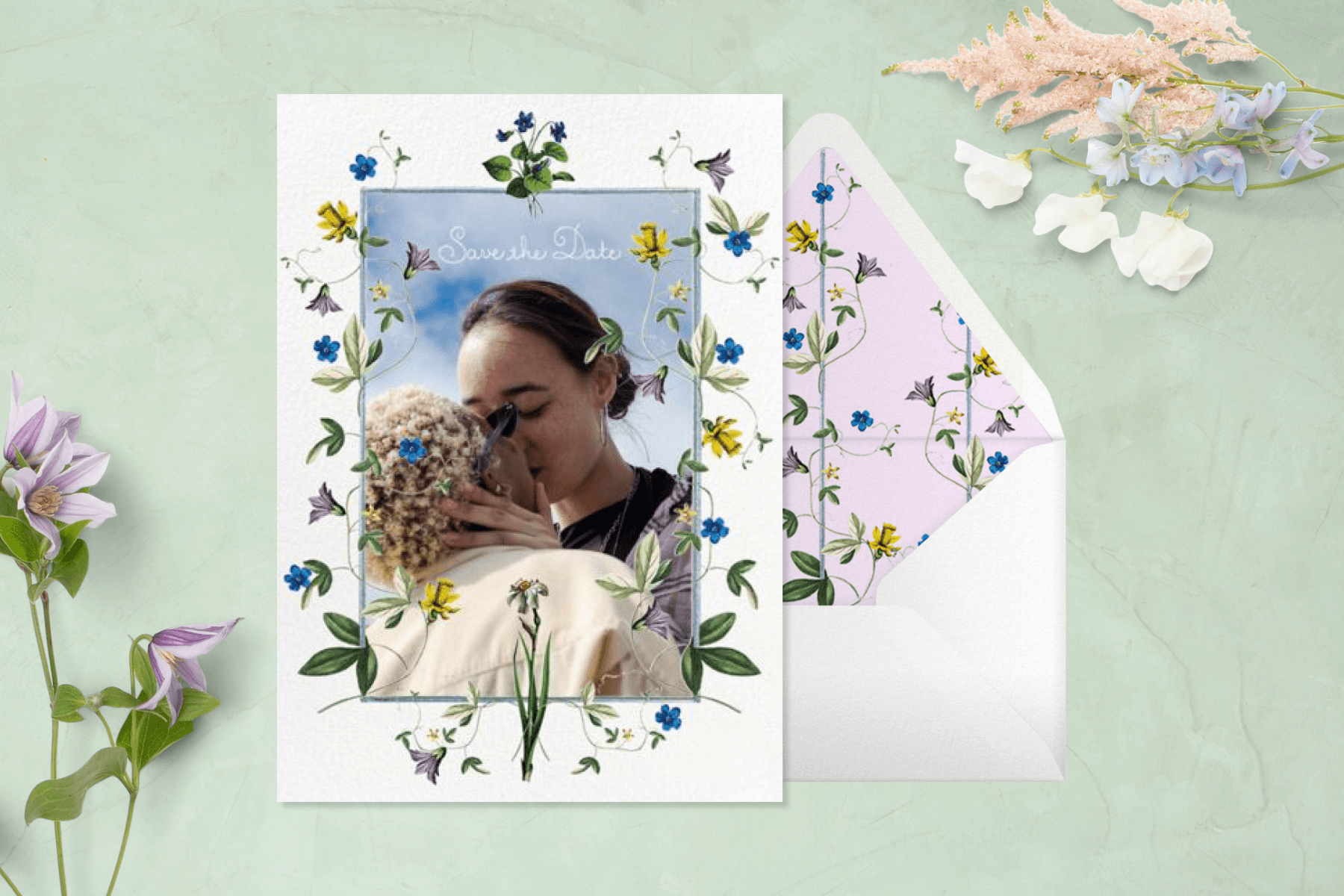 A vertical save the date with a photo of two people kissing with a border of delicate flowers and vines. The background is a pastel green with pastel flowers laid out on either side and a matching envelope.