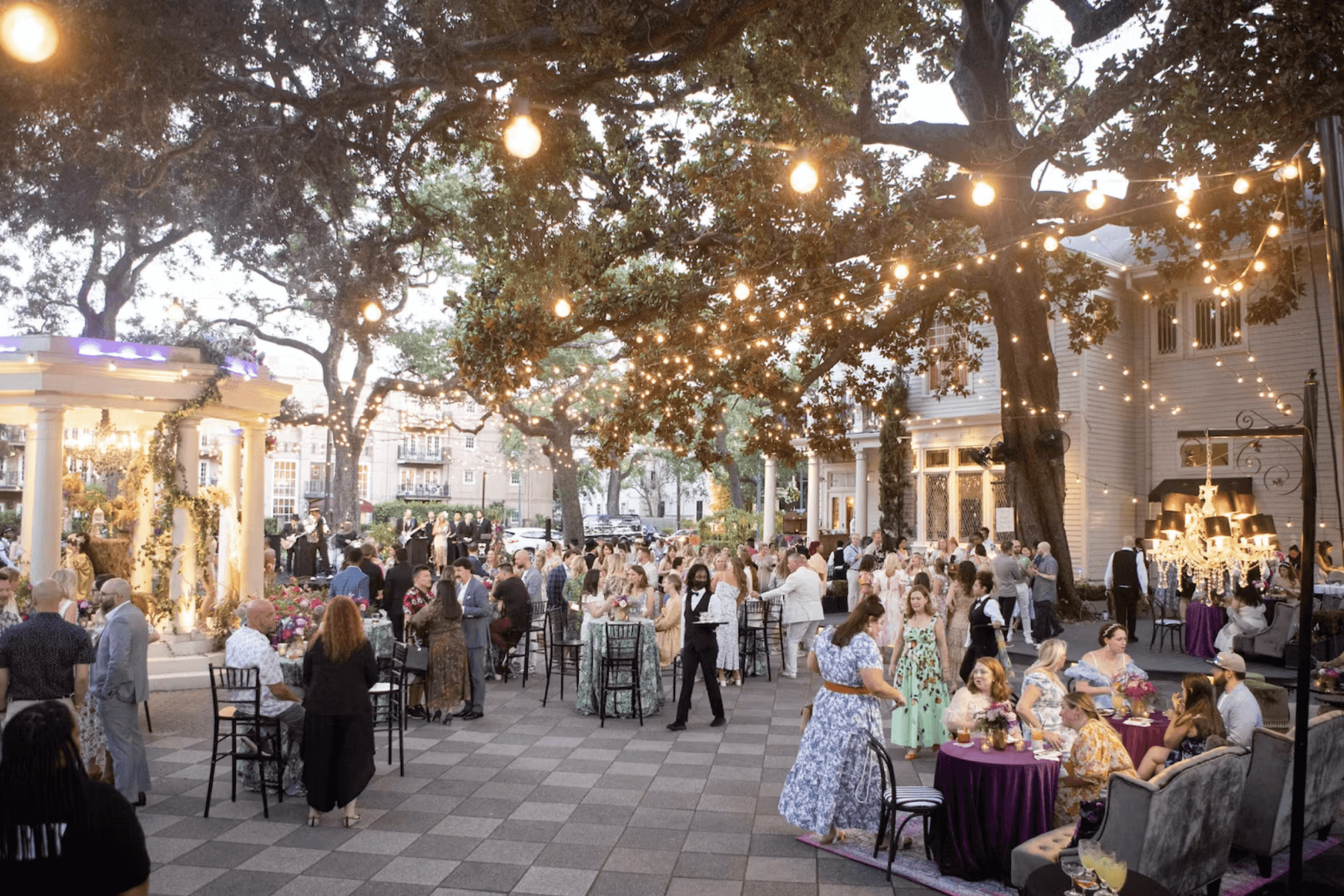 Many guests are pictured at an outdoor event beneath lighted trees.