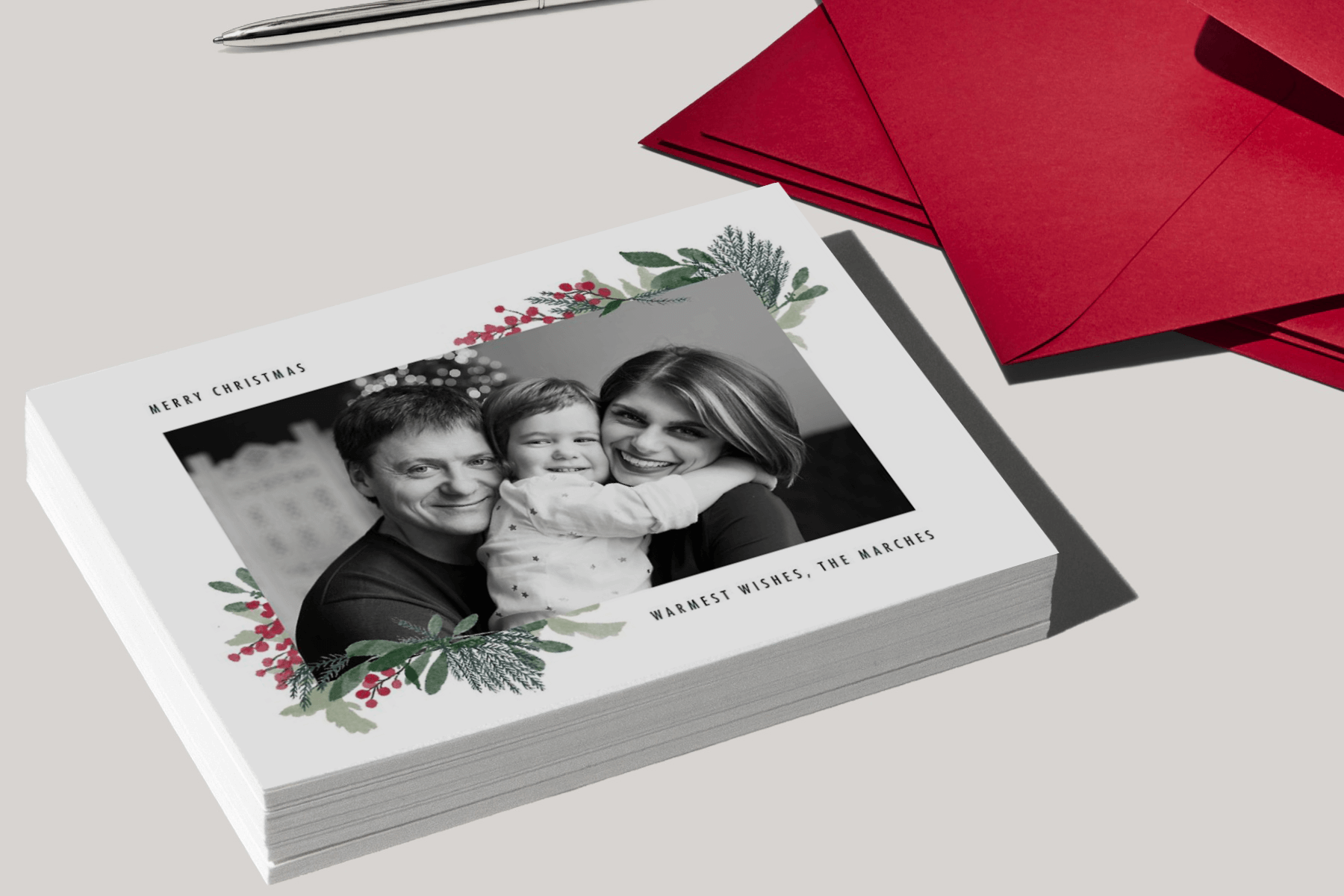 A stack of holiday cards with a black and white photo of a family surrounded by holly sprigs, next to several red envelopes.