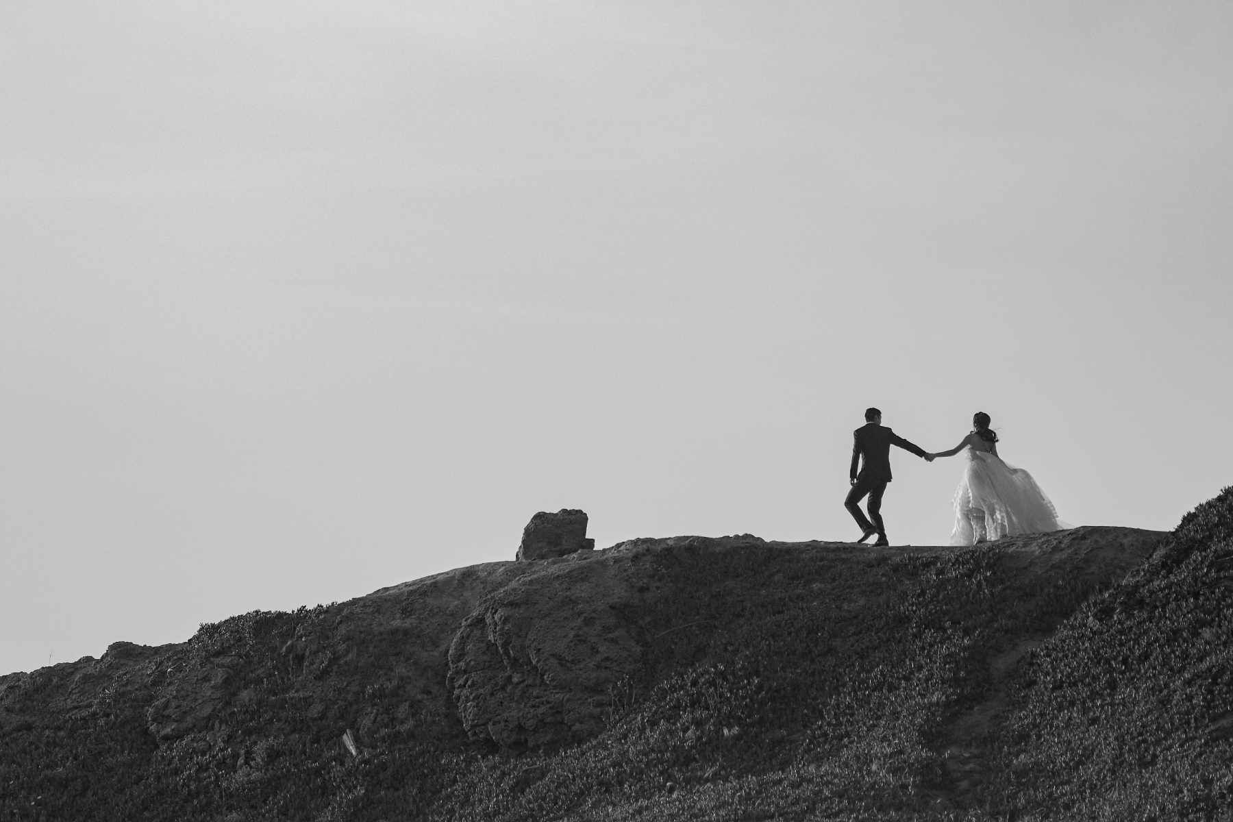 ALT: A black-and-white photo of a bride and groom holding hands and walking along a cliff.