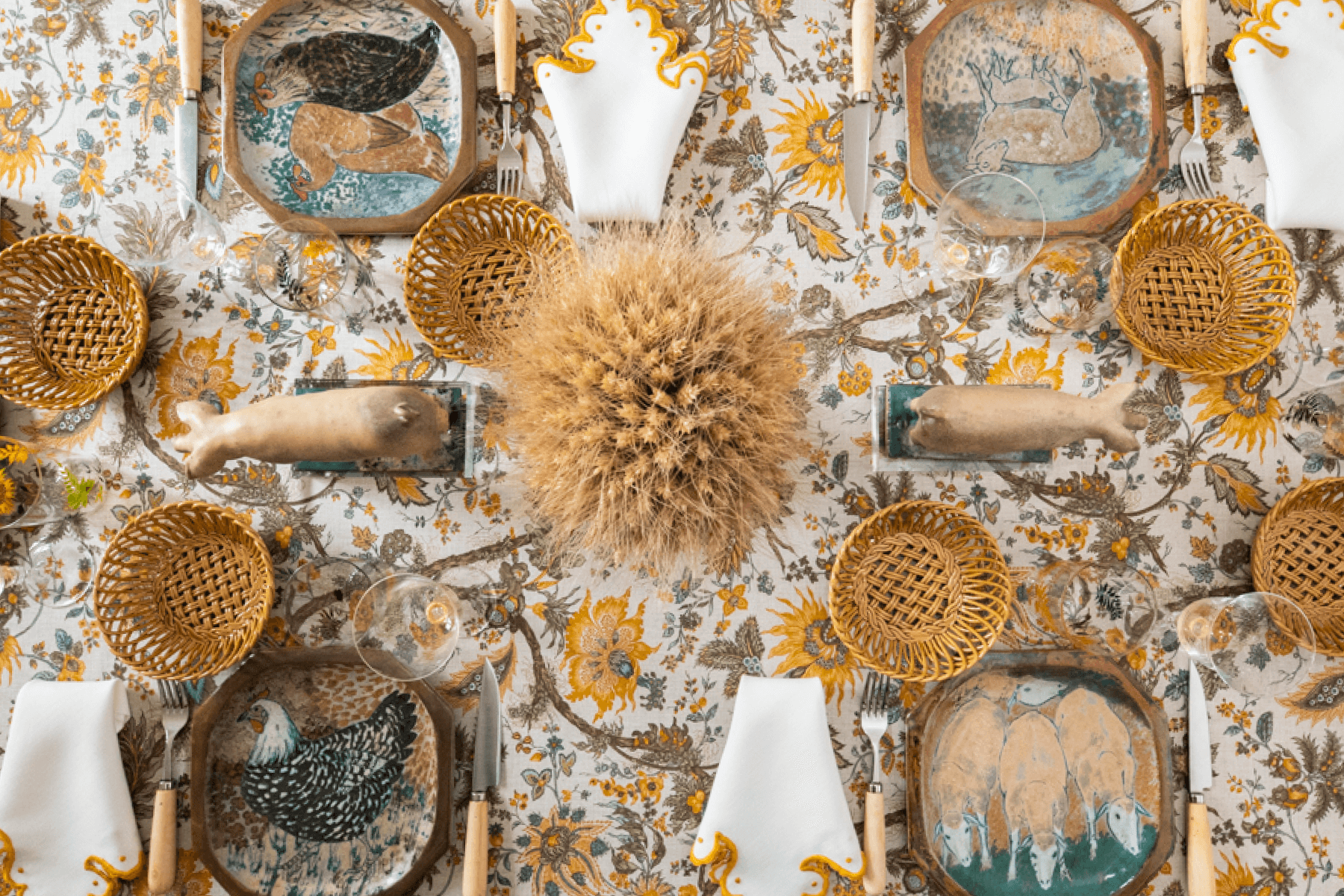 Wide crop of a dinner table set with a yellow floral tablecloth, ceramic plates featuring farm animal designs, straw baskets, and wheat accents. 