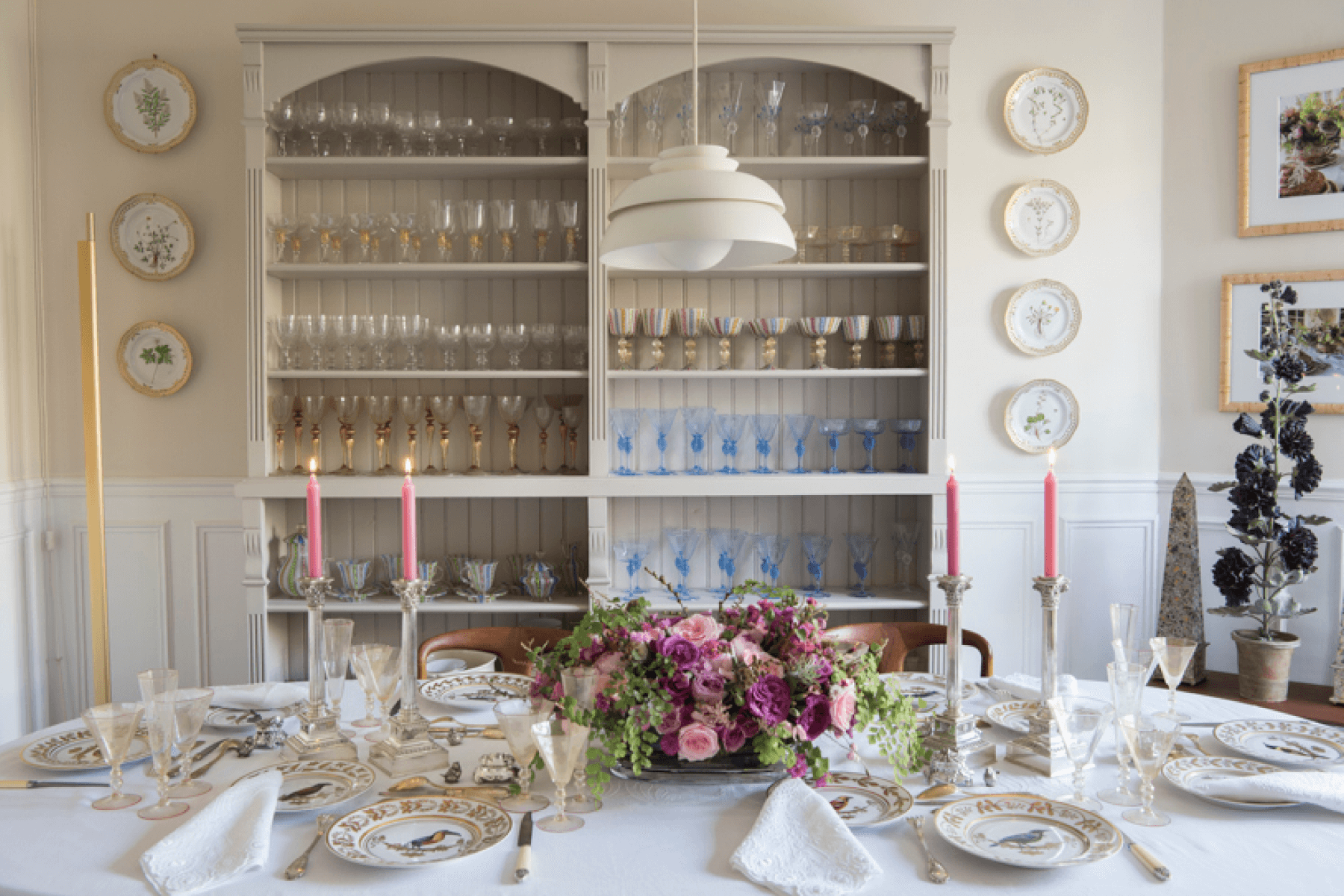 Wide crop of a white dinner table set with gold-accented tableware and pink candles, featuring a pink and purple flower arrangement. 