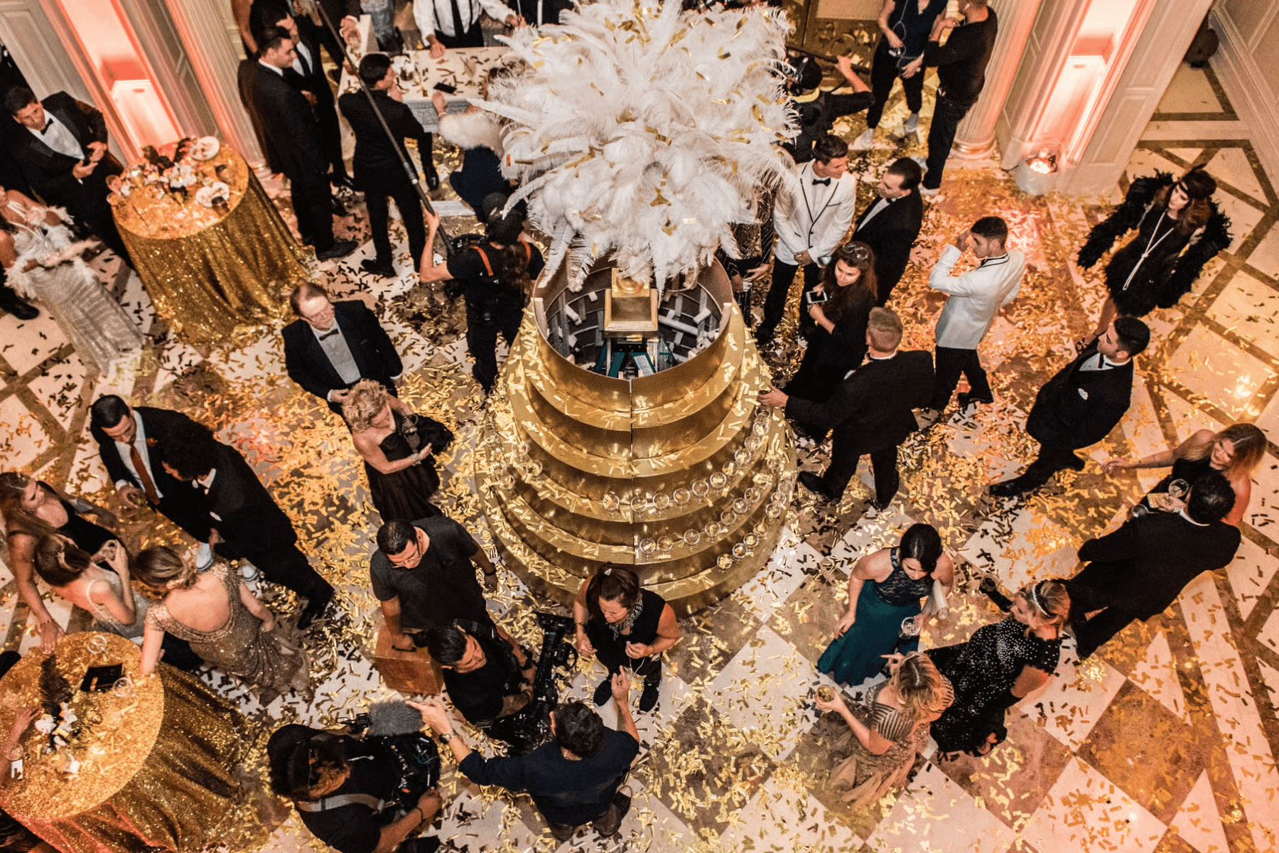A formal party viewed from above with gold tiered tables, white ostrich feathers, marble checkerboard floors, and party goers in black tie outfits.