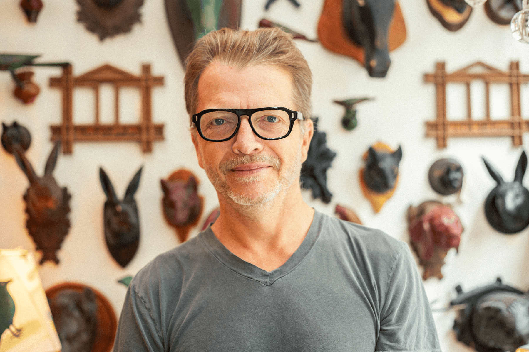 Portrait of John Derian inside his studio wearing a grey t-shirt and thick black glasses, standing in front of a wall of ceramic and wood animal sculptures.