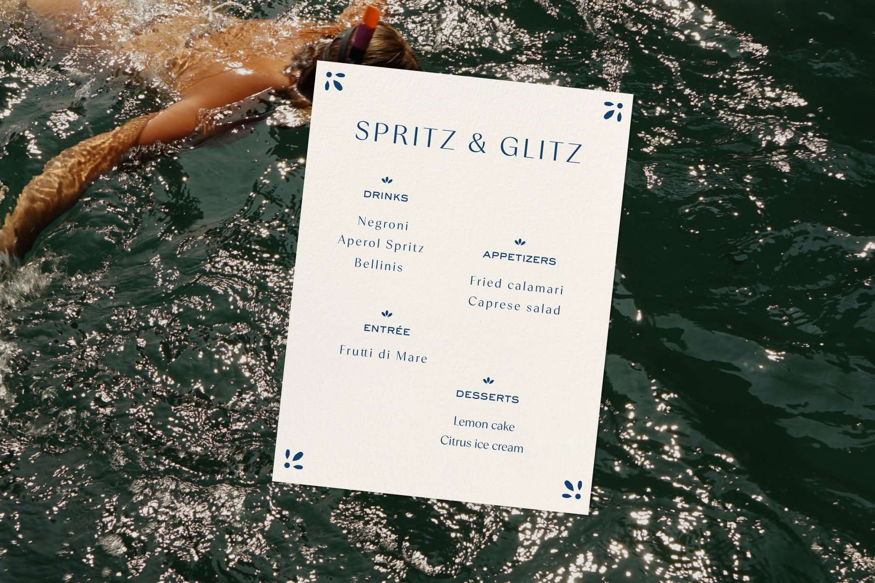 ALT: A white invitation with dark blue text reading ‘Spritz & Glitz’ along with a menu listing drinks, appetizers, an entrée, and desserts, over a backdrop image of a body of water and a person snorkeling.