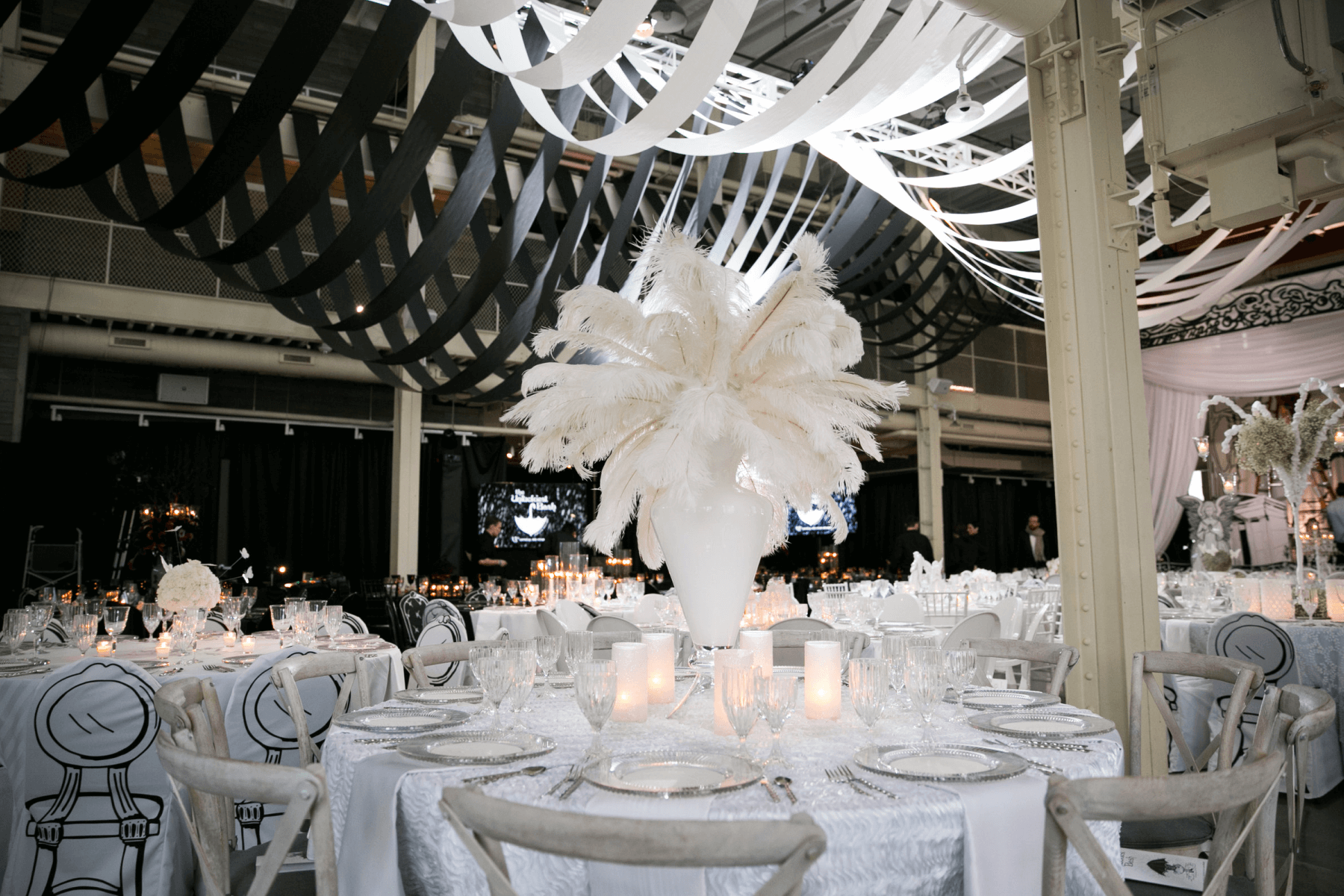 A large event space with round white tables and chairs, white ostrich feathers as centerpieces, and white and black fabric strips draped from the ceiling.