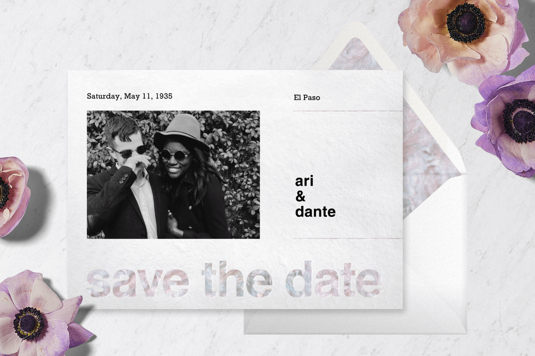 A save the date card with a black and white photo of a couple on the left and mother of pearl-patterned text lies beside purple anemone flowers.