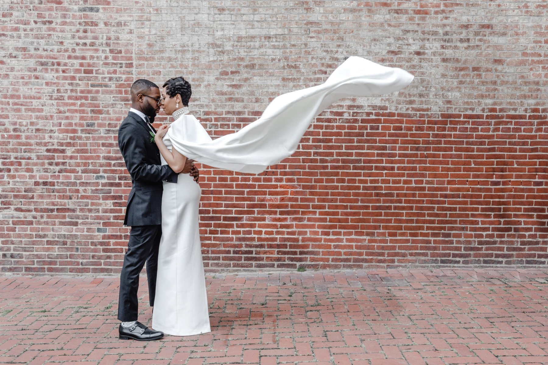 A wedding couple stands in front of a brick wall. The bride’s cape billows in the wind.