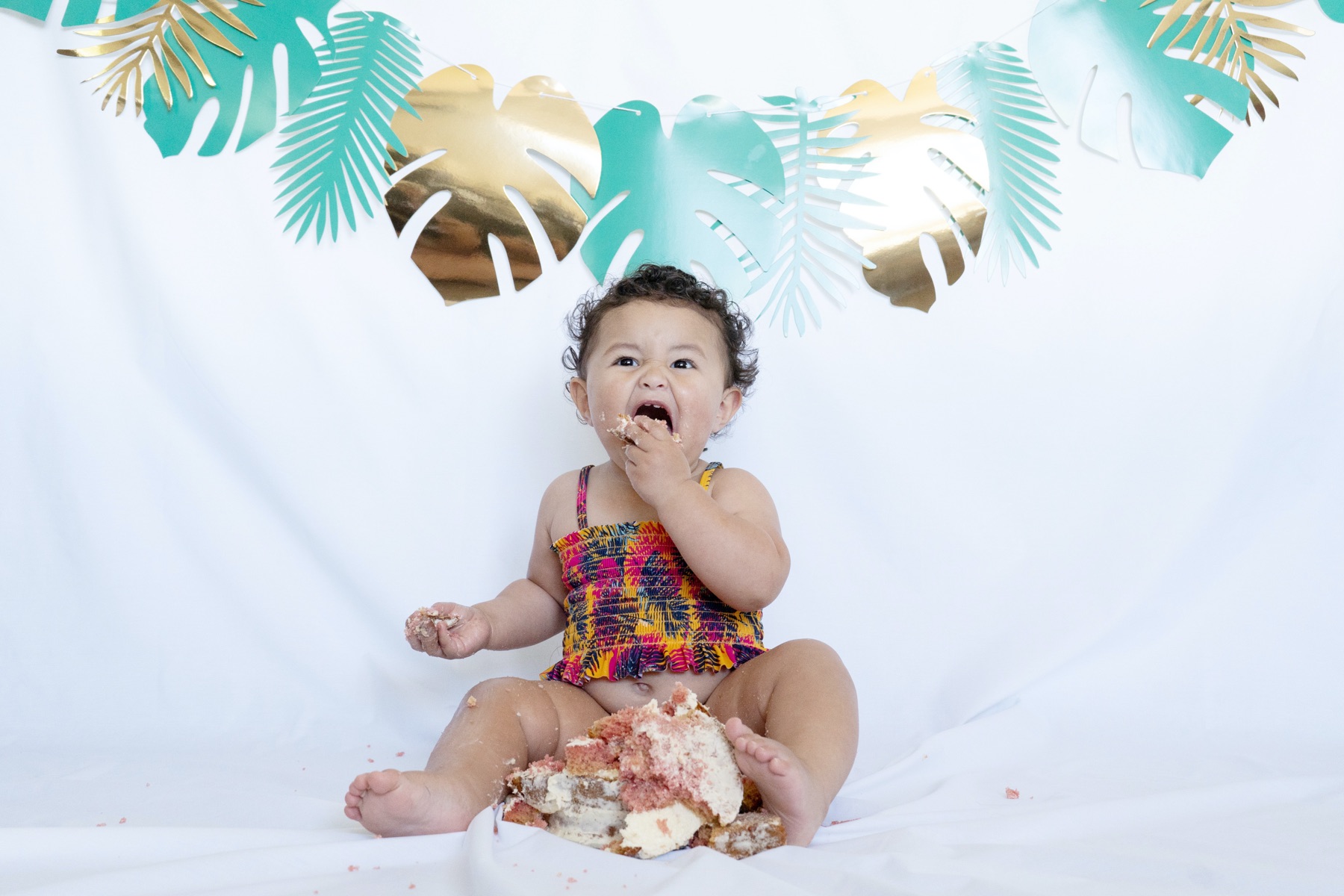 Photo of a baby eating a cupcake in front of a banner of monstera fronds.