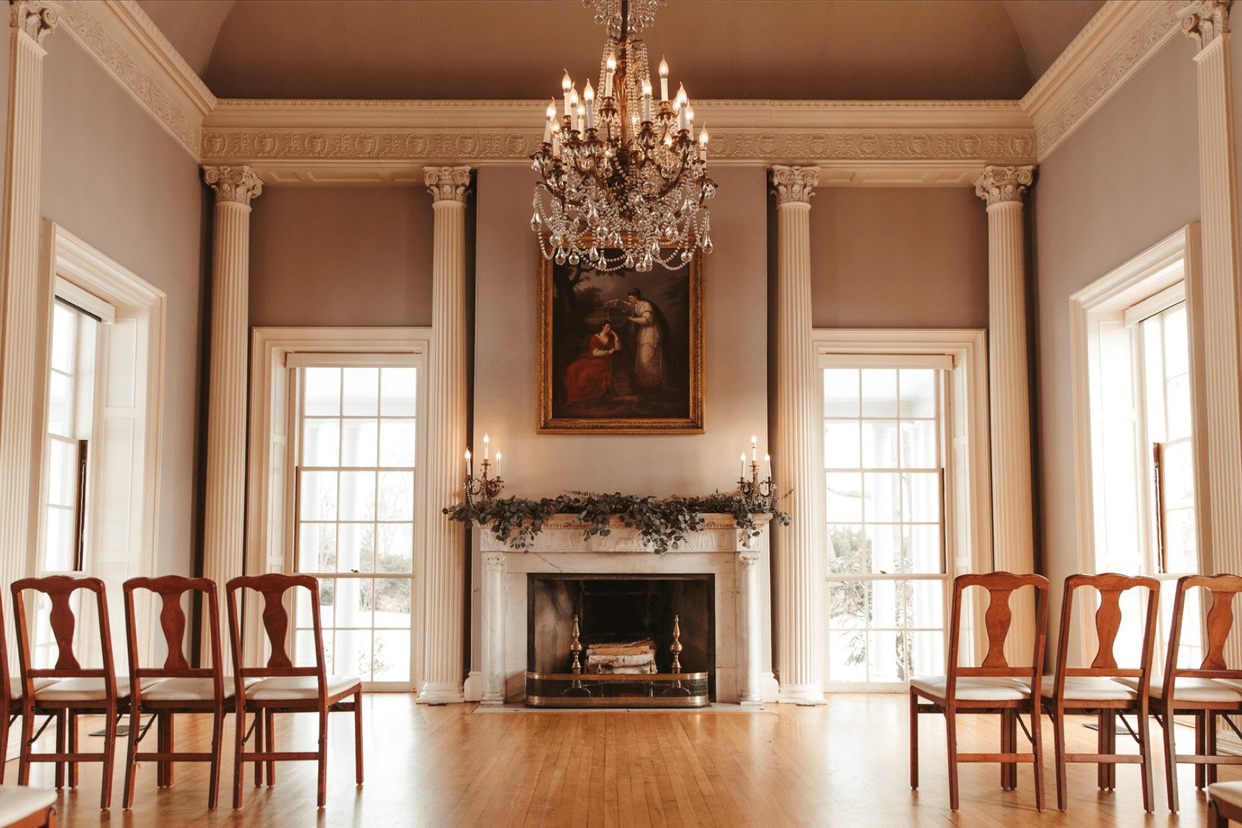 A large room with columns, fireplace, and a chandelier with chairs set up for wedding guests.