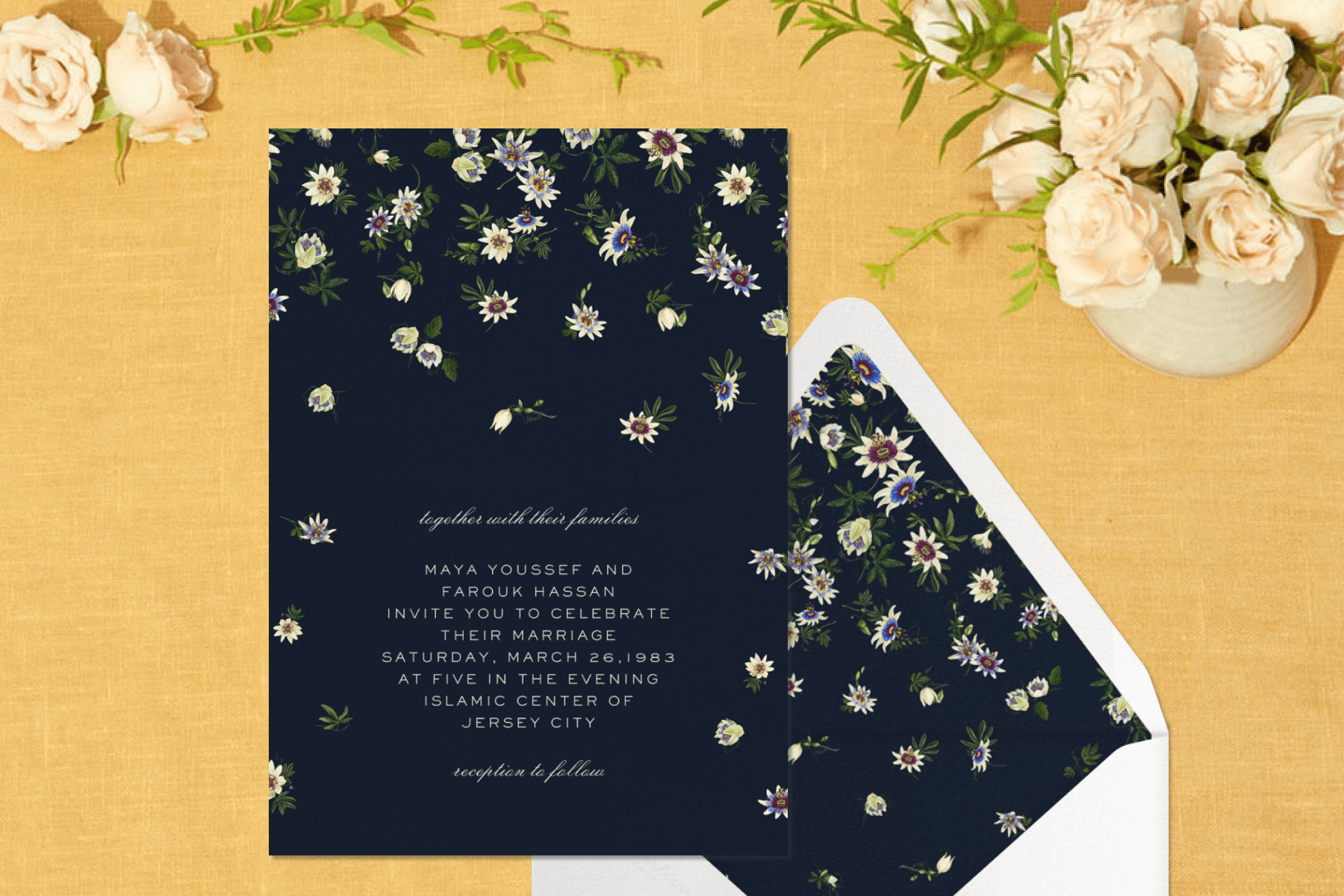 A navy wedding invitation with passion flower designs, paired with a matching envelope liner. The card is propped with roses and a yellow background.