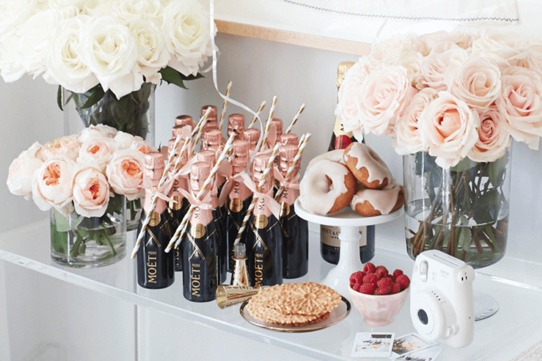 A shelf with white and pink roses, mini Champagne bottles, doughnuts, and a white Fuji Instax camera.