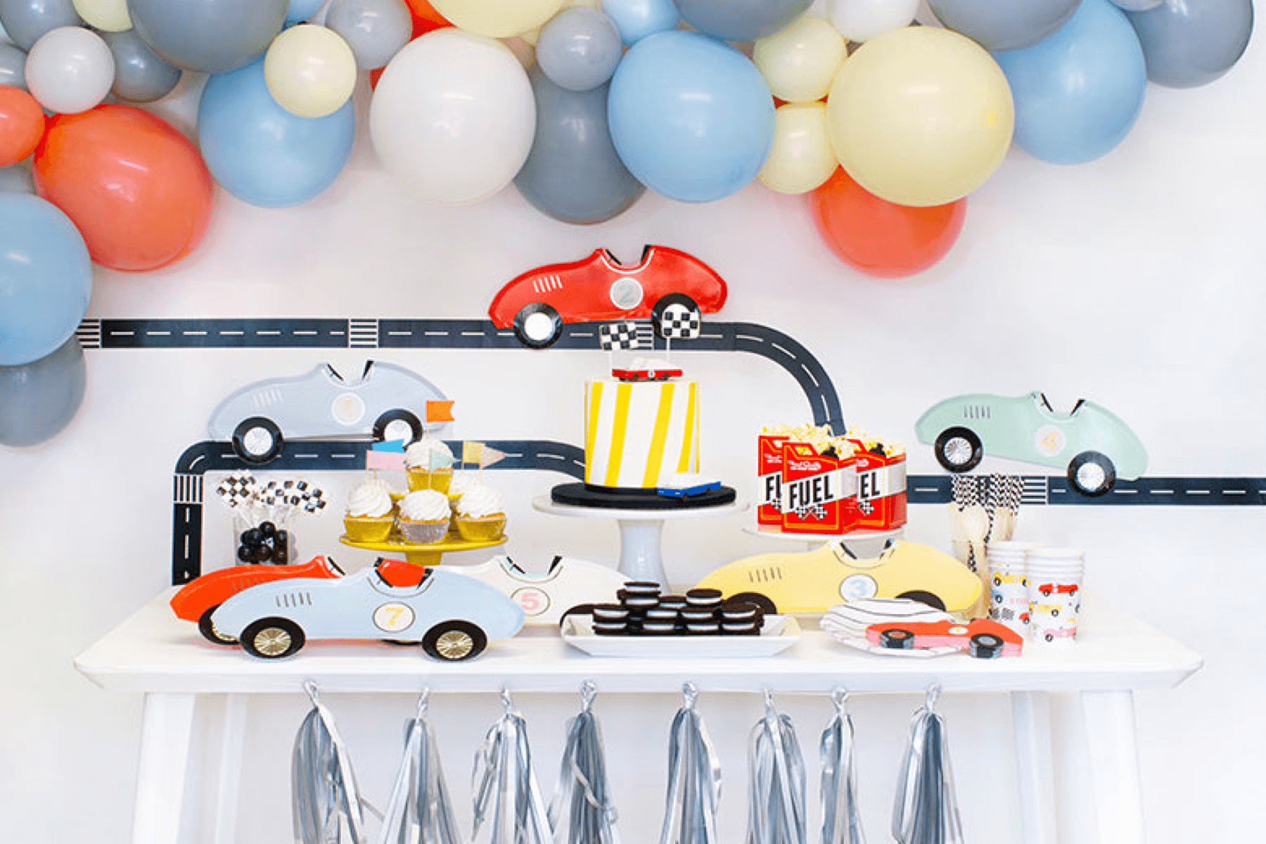 A party table set with race car-shaped plates, a striped cake, cupcakes, and multicolored balloons. 