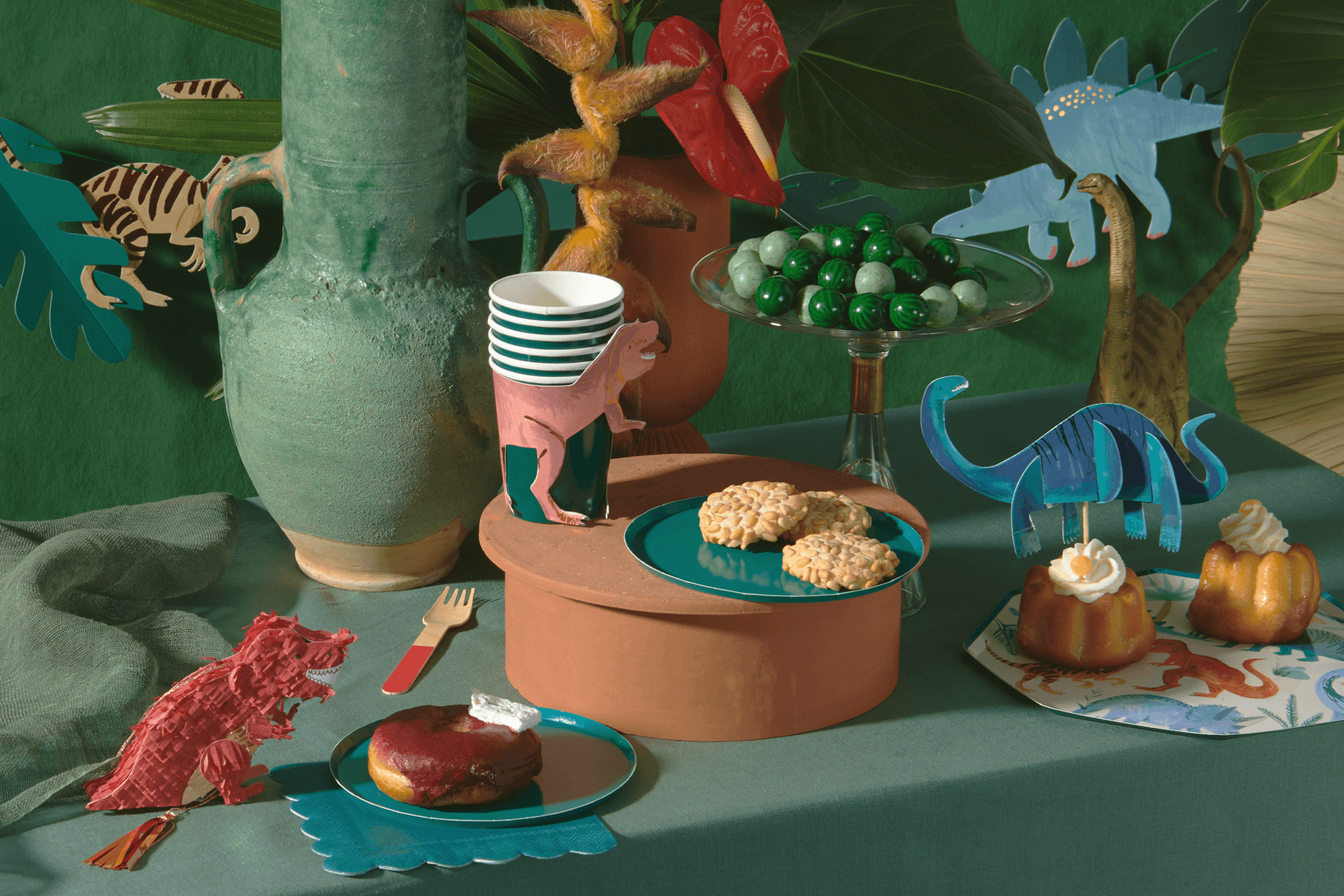 Party scene featuring desserts and dinosaur-themed tableware lie cups and cardboard cutouts.