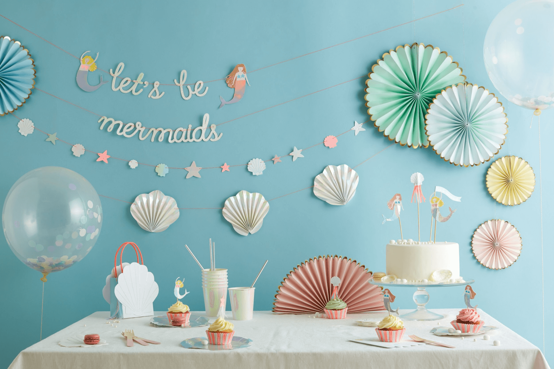 A party scene shows a table top and wall decorated with mermaid and under-the-sea themed party supplies including banners, honeycomb decor, balloons, and cake toppers.