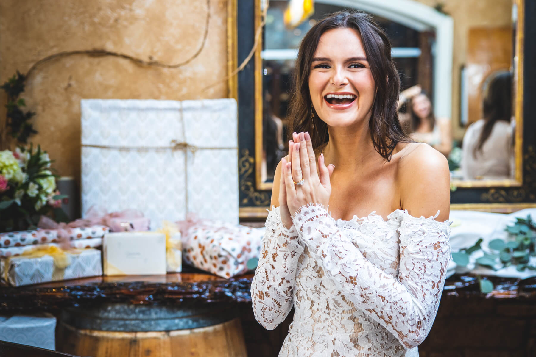 Photo of a happy bride wearing a white lace top in front of a stack of presents.