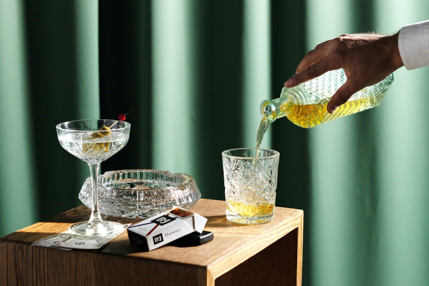 Image of a hand pouring a drink into a crystal glass with another cocktail and cigarettes on the table in front of a green background.