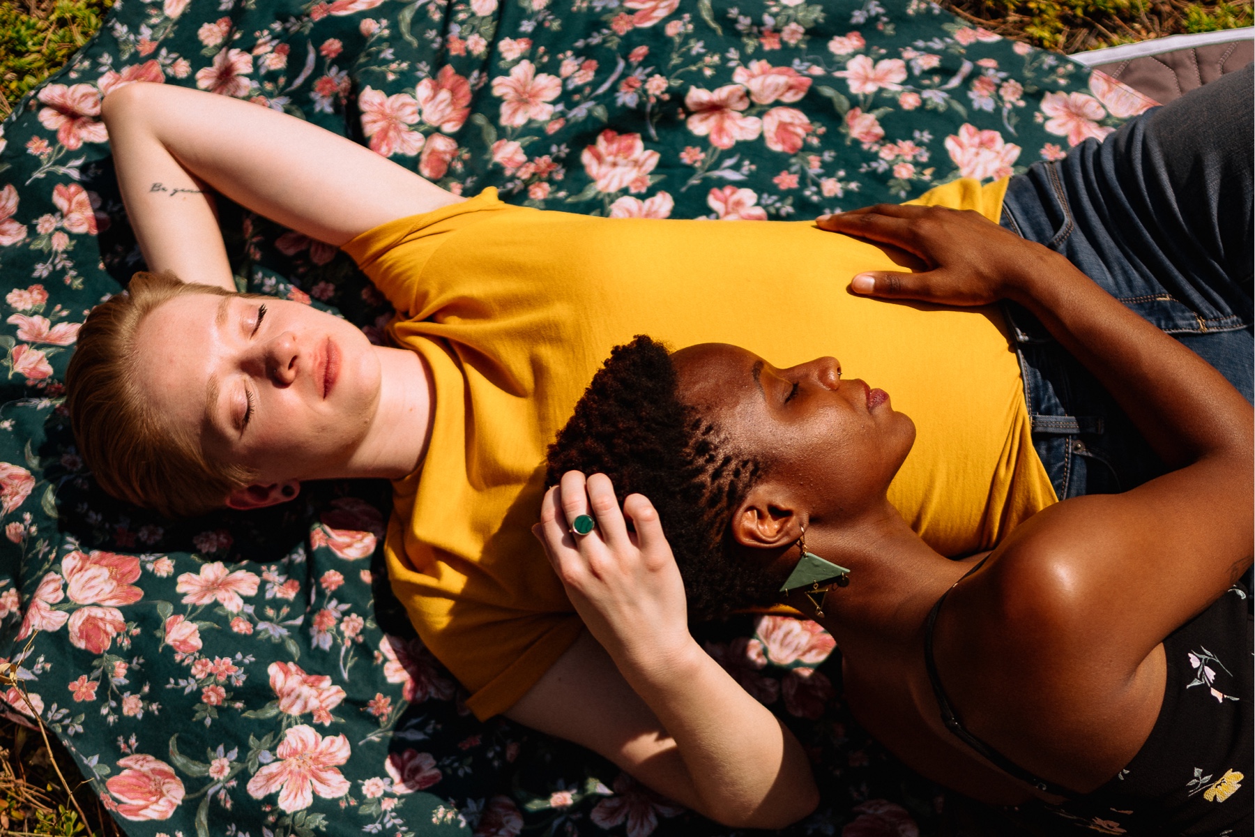 Two women posing together for a couples' photoshoot, one is laying her head on the other's stomach. Background is muted florals.