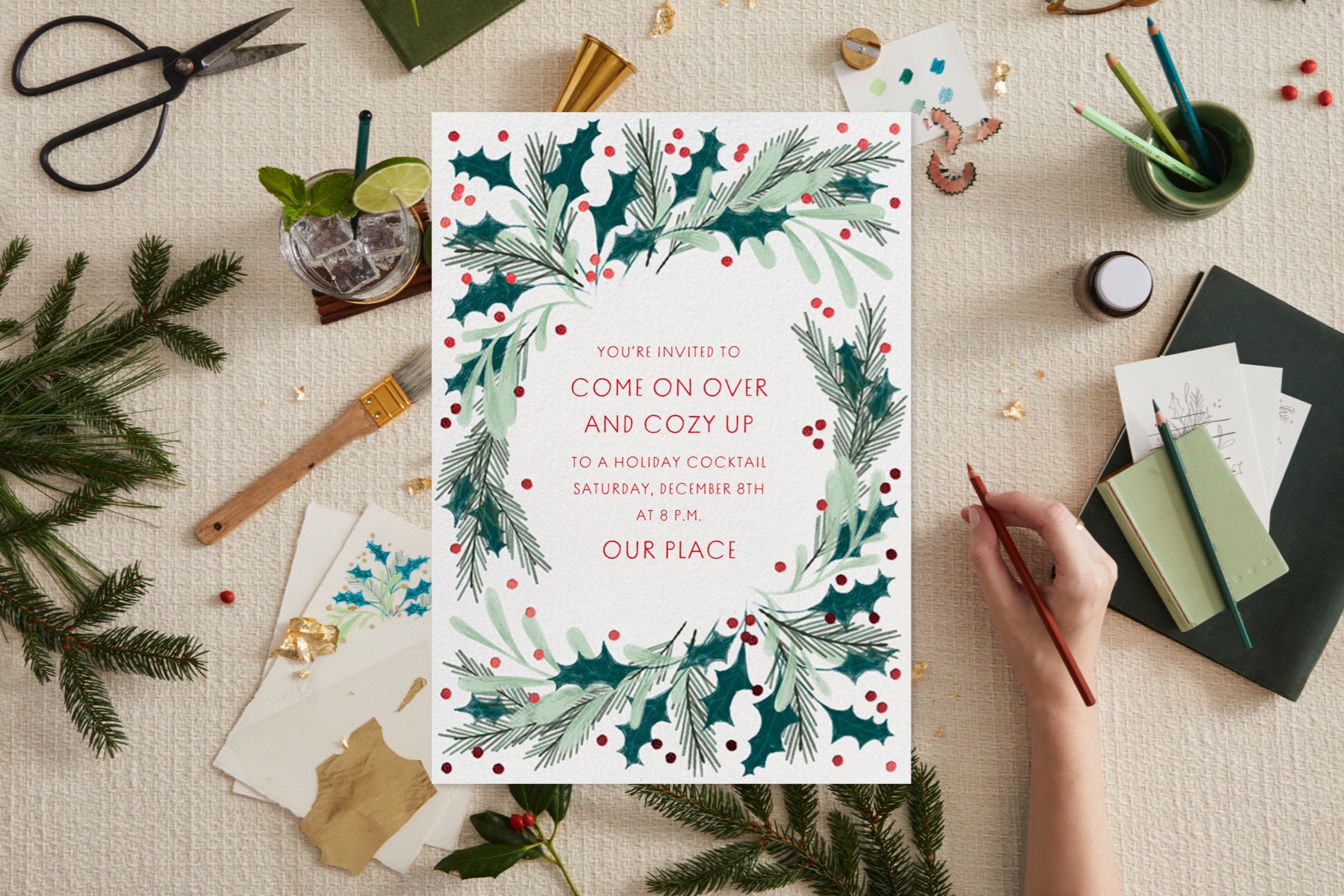 A holiday invitation featuring a wreath design surrounded by a scene with the designer’s inspiration items, like greenery, gold foil, and colored pencils. 