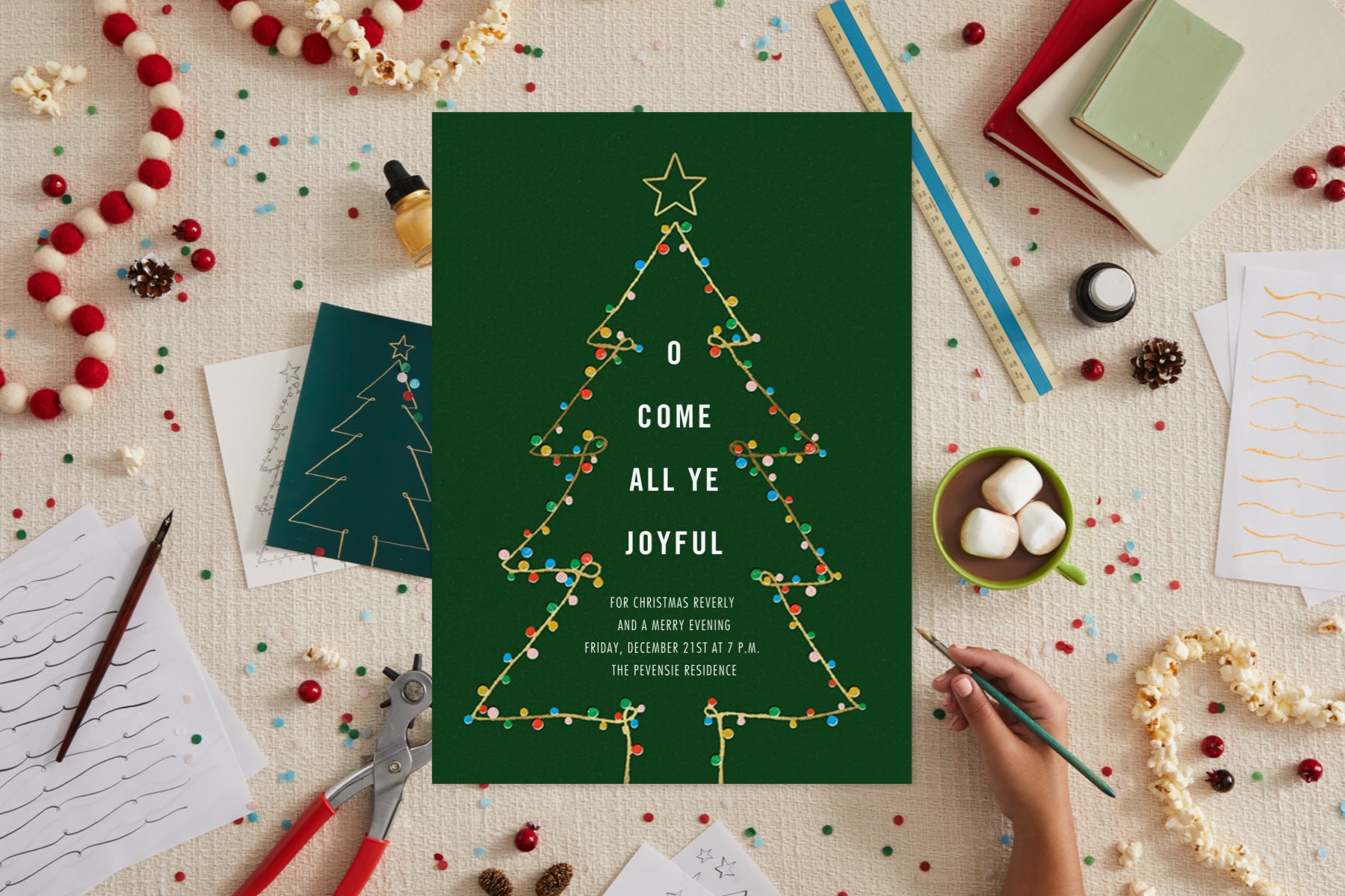 A Christmas invitation surrounded by objects of inspiration like sketch paper, garland, and hot cocoa.
