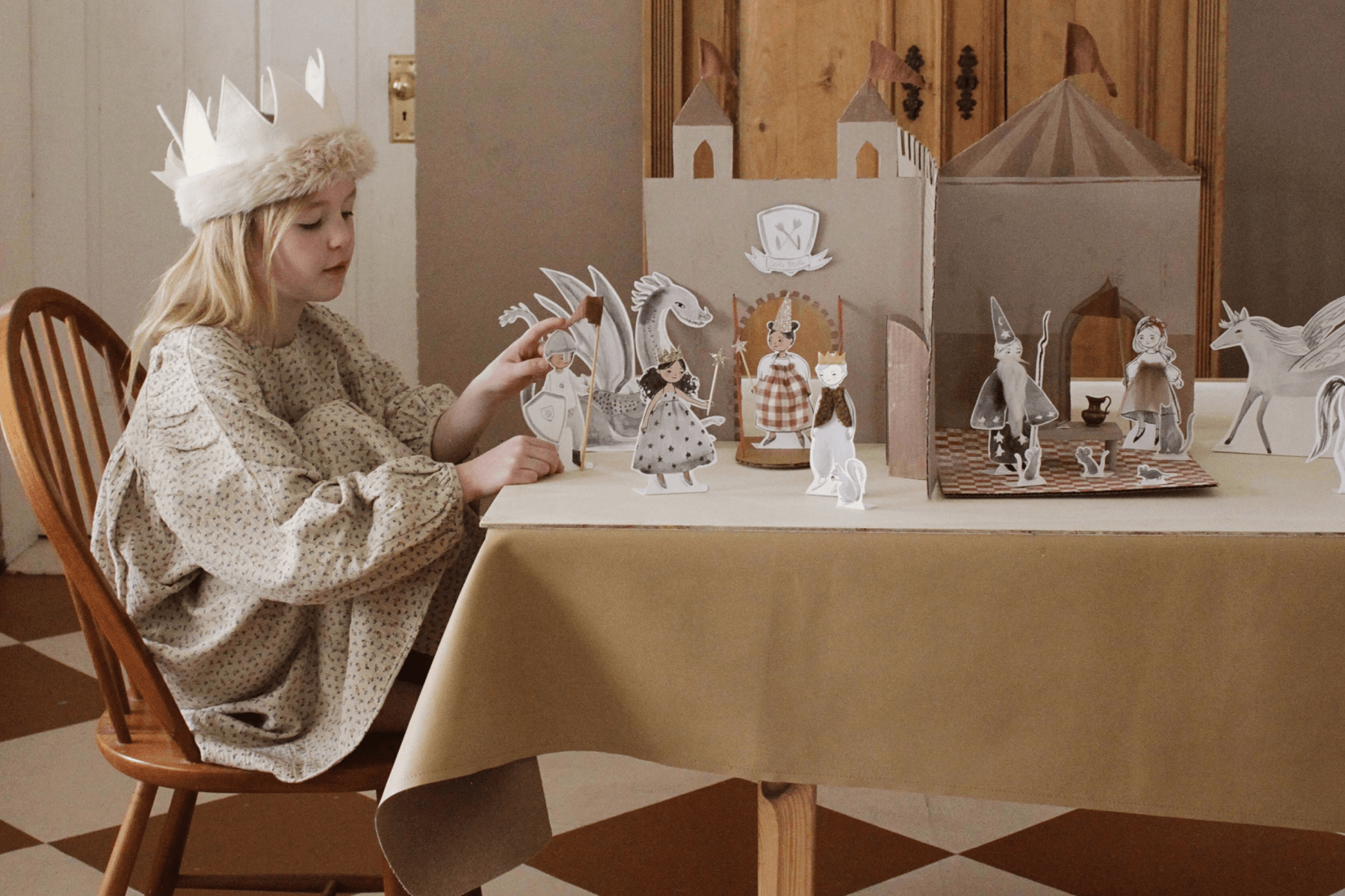 Young girl in a paper crown sits at a kitchen table and plays with a paper cutout castle and illustrated paper dolls.