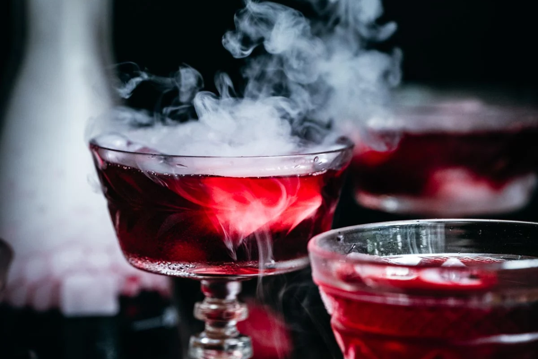 A close-up photograph of dark red cocktails with dry ice smoke billowing out.