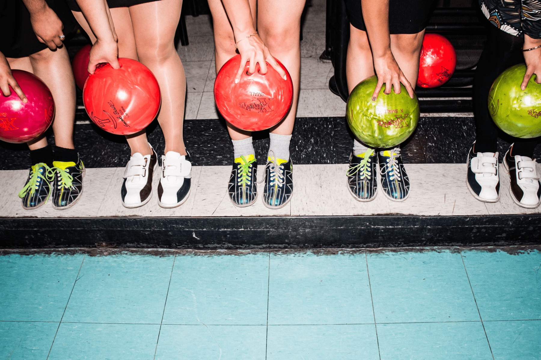 The legs of people wearing bowling shoes and holding colorful bowling balls.